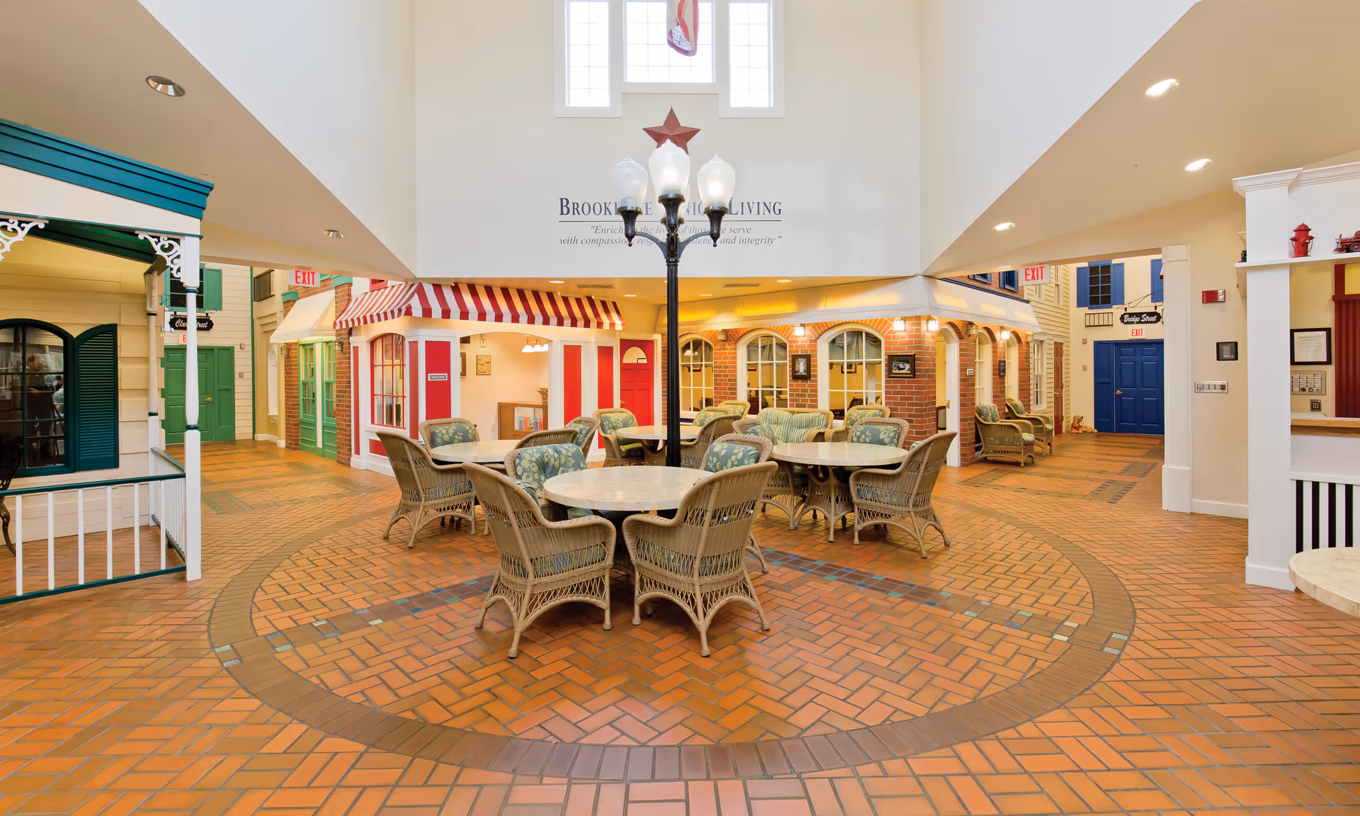 Interior common area of Brookdale Troutdale featuring round tables with wicker chairs arranged on a brick-patterned floor. The space is designed like a small village street with colorful storefront facades, including green, red, and blue doors and windows. A tall lamp post stands in the center, and large windows above let in natural light.