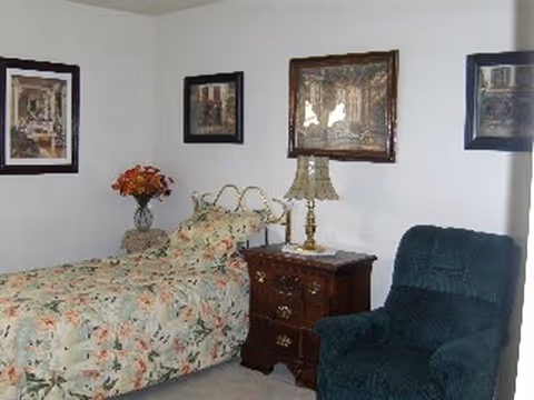 A small bedroom with a single bed covered in a floral bedspread, a wooden nightstand with a decorative lamp, a vase with flowers, three framed pictures on the walls, and a dark green upholstered armchair.