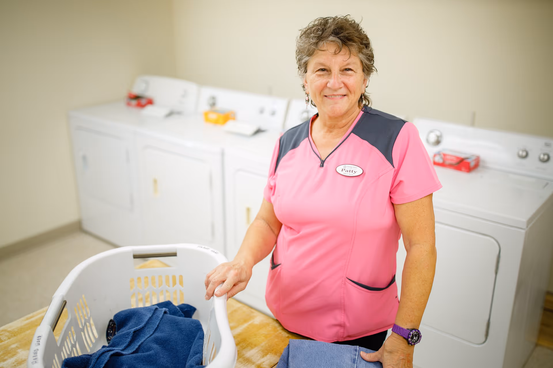 A woman wearing a pink and gray uniform with a name tag that says Patty stands in a laundry room. She is smiling and holding a laundry basket with folded clothes. Behind her are white washing machines and dryers against a beige wall.