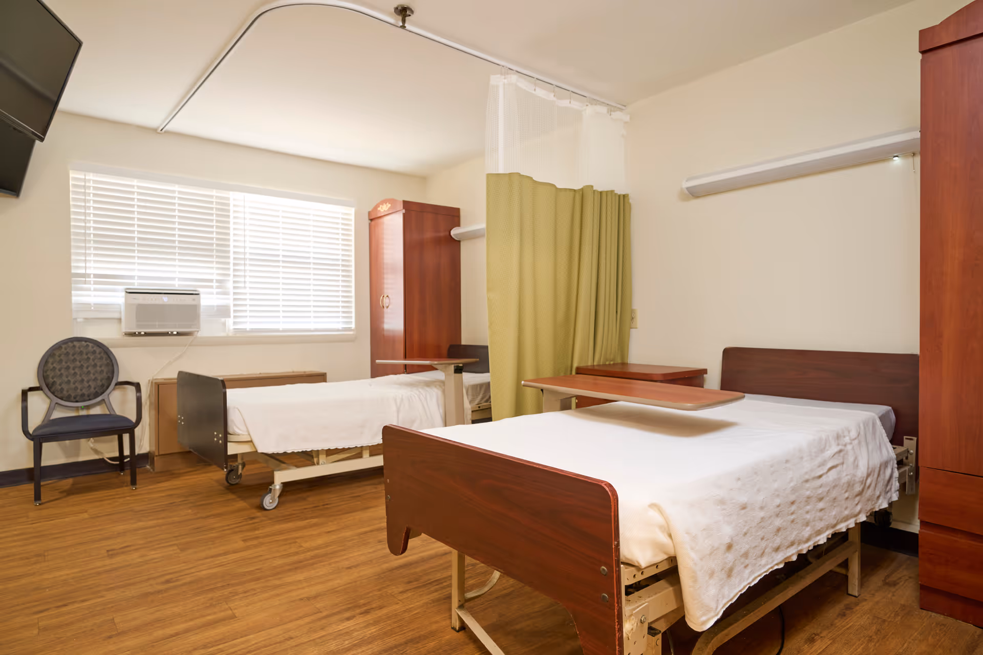 A senior care room with two hospital-style beds, wooden furniture, a privacy curtain, and a window with blinds.