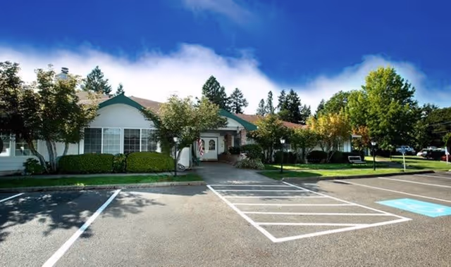 Exterior view of a single-story building with a green roof and white walls, surrounded by trees and bushes, with a parking lot in front including a handicapped parking space.