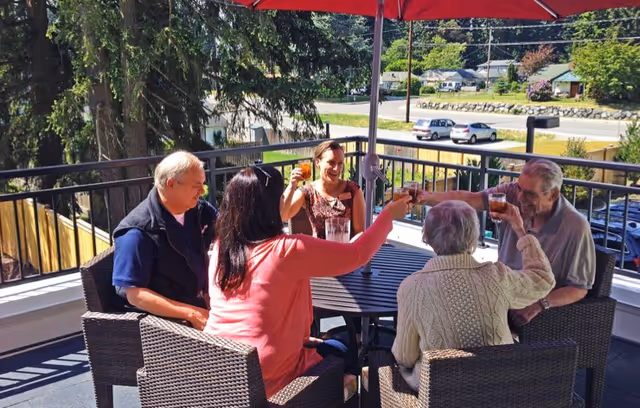 Five people sitting around a round outdoor table on a patio, raising their glasses in a toast under a large red umbrella. The background shows trees, a road, parked cars, and houses.