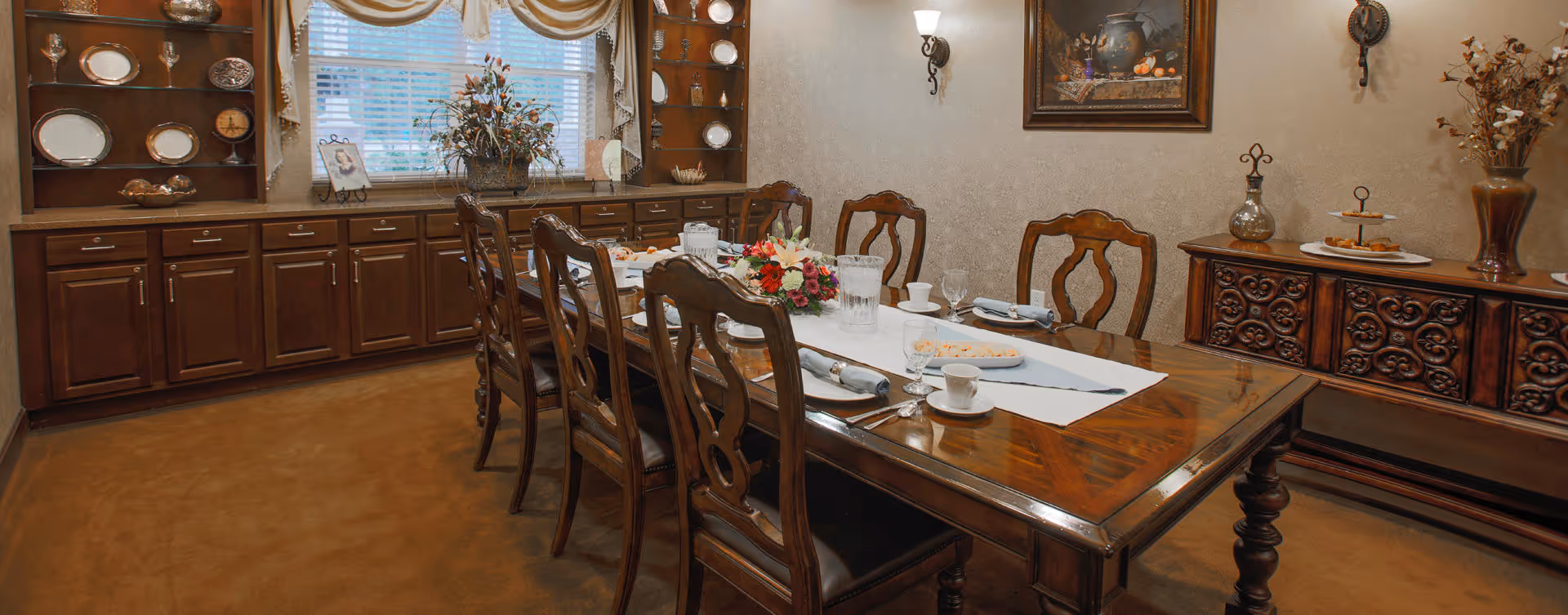 A traditional dining room with a long wooden table set for a meal, surrounded by eight wooden chairs. The table has a white runner, plates, cups, glasses, and a floral centerpiece. The room features wooden cabinetry with decorative plates and bowls, a window with draped curtains, wall sconces, a framed painting, and a sideboard with decorative items and dried flowers.