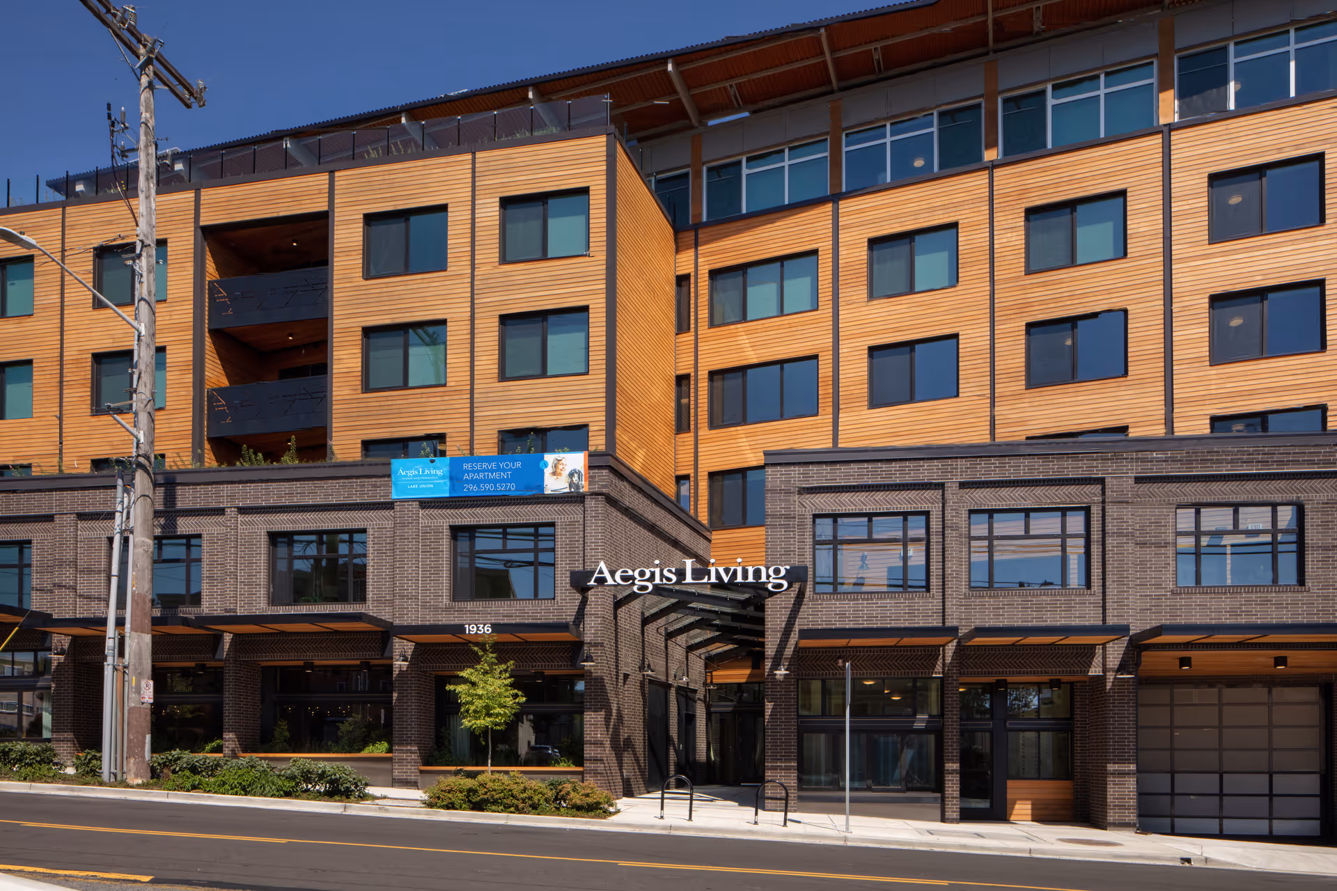 Exterior view of Aegis Living Lake Union, a multi-story senior living facility with a modern design featuring wood paneling and large windows. The building has a brick lower facade and a covered entrance with the name 'Aegis Living' displayed above it. There is a street and sidewalk in front of the building with some greenery and a utility pole on the left side.