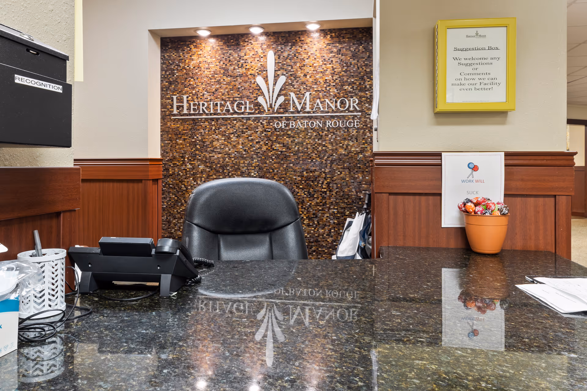 Reception desk area at Heritage Manor of Baton Rouge with a black office chair, a telephone, a pen holder, a suggestion box on the wall, and a small pot of lollipops on the counter. The background features a mosaic tile wall with the Heritage Manor of Baton Rouge logo.