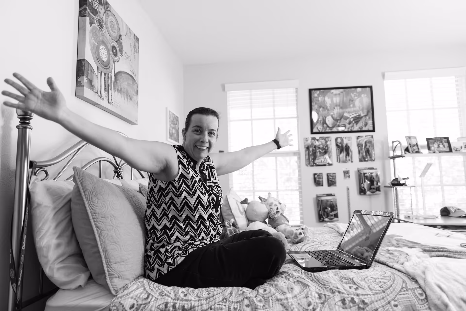 A person sitting cross-legged on a bed with arms outstretched, smiling at the camera. The bed has patterned bedding and several pillows. A laptop is open on the bed. The room has two large windows with blinds, and the walls are decorated with various framed pictures and artwork.