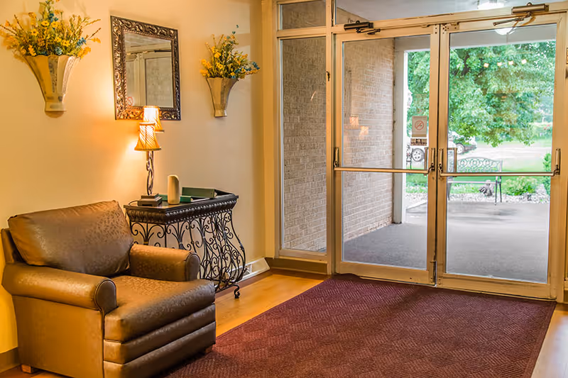 A cozy entrance area with a brown leather armchair, a decorative black metal console table with books and a lamp, two wall-mounted flower arrangements with yellow flowers, a framed mirror, and glass double doors leading outside to a green garden area.