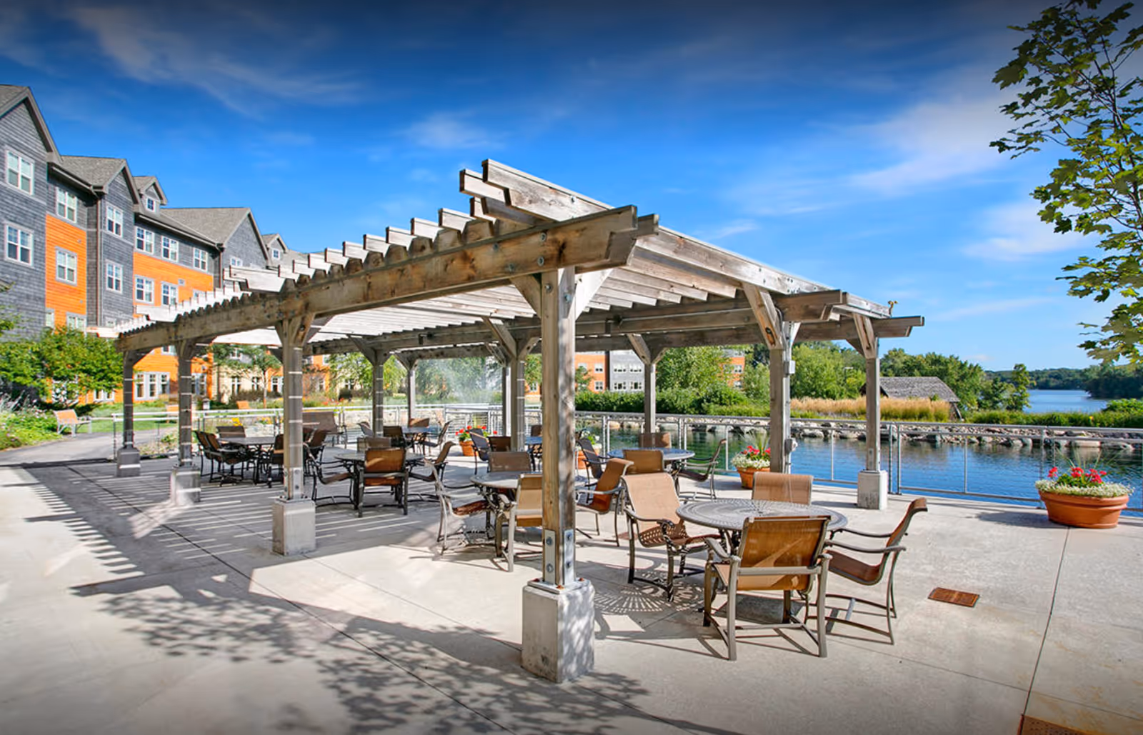 Outdoor patio area at Johanna Shores featuring a wooden pergola with multiple round tables and chairs arranged underneath. The patio overlooks a serene body of water with greenery and a clear blue sky in the background. The building of the facility is visible on the left side.