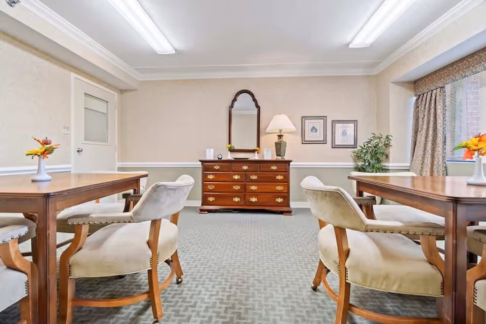 A well-lit room with two wooden tables, each surrounded by upholstered chairs with wooden legs. At the far end of the room is a wooden dresser with brass handles, topped with a table lamp, a mirror, and some small decorative items. The walls are light-colored with a chair rail molding, and there are two framed pictures hanging above the dresser. A window with patterned curtains is on the right side, and a potted plant is placed near the window.