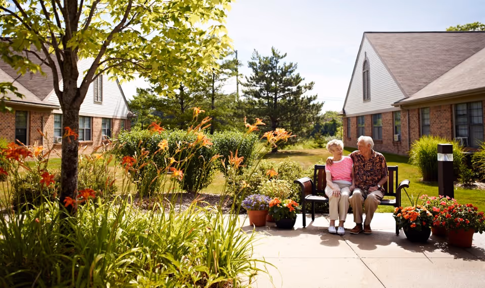 An elderly couple sitting on a bench outside in a garden area surrounded by flowering plants and greenery, with two brick buildings in the background under a sunny sky.