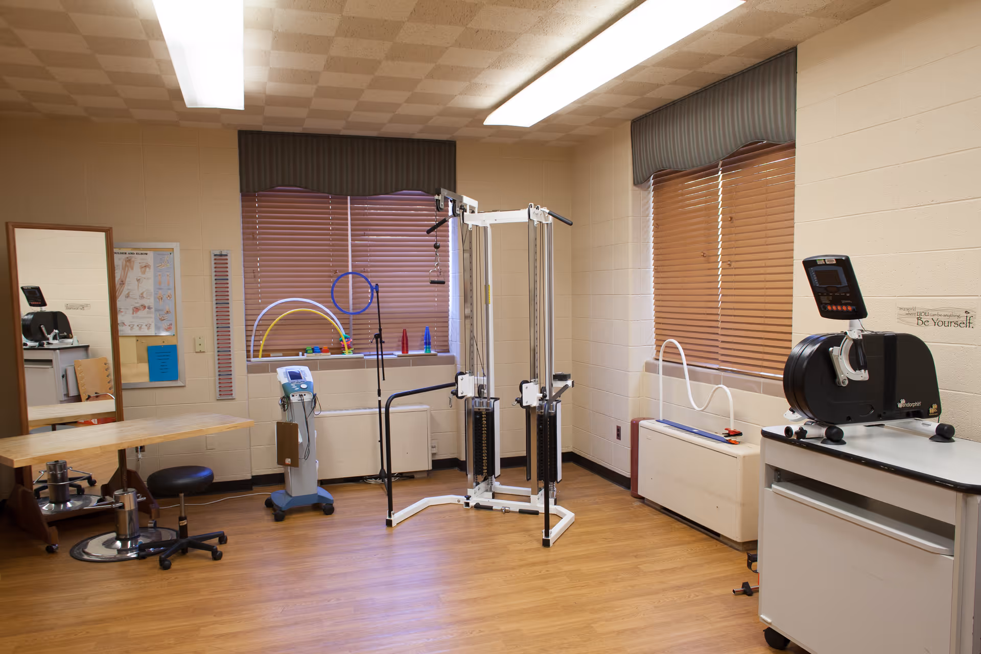 A therapy/exercise room with weight machines, rehab equipment, and a small desk against beige walls and wood flooring.