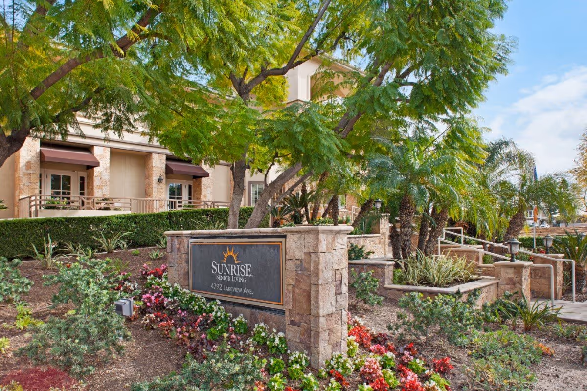 Stone entry sign that reads 'Sunrise Senior Living' at the landscaped front of the facility with trees, flowers, and the building behind it.