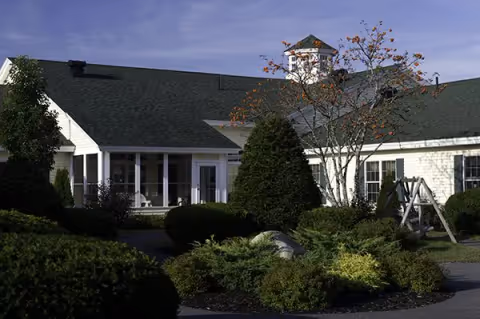 Front exterior of a single-story memory care building with a green roof, landscaped shrubs, a small porch, and a cupola.