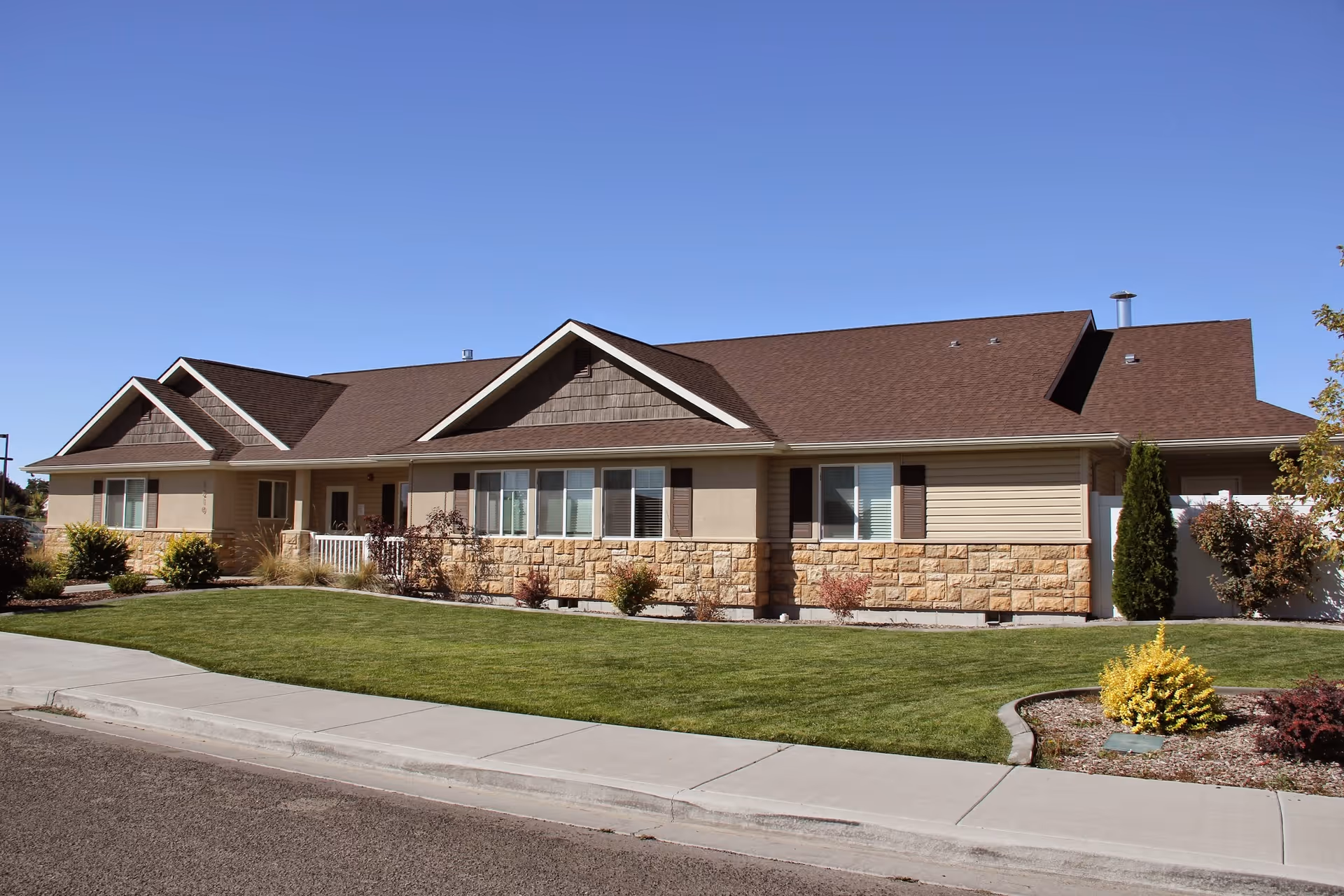 Single-story residential-style building with a brown roof, beige siding, and stone accents. The front yard has a well-maintained green lawn, small shrubs, and a concrete sidewalk. The sky is clear and blue.