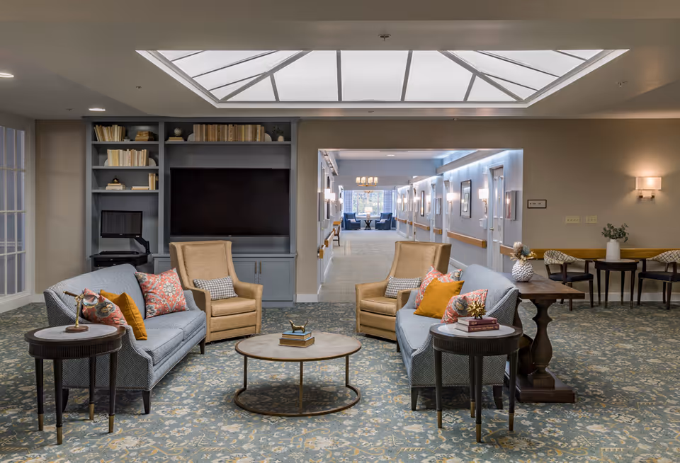 A cozy and well-lit living room area in a senior living facility featuring two blue patterned sofas with colorful throw pillows, two beige armchairs, a round coffee table with books, and side tables with decorative items. Behind the seating area is a built-in bookshelf with books and a large black screen. The room has a large skylight, patterned carpet, and a hallway leading to another seating area with windows in the background.