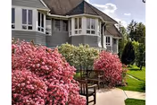 Exterior view of a senior living facility building with gray siding and multiple windows. In front, there are vibrant pink flowering bushes and a wooden bench on a paved walkway. Green grass and trees are visible in the background under a partly cloudy sky.