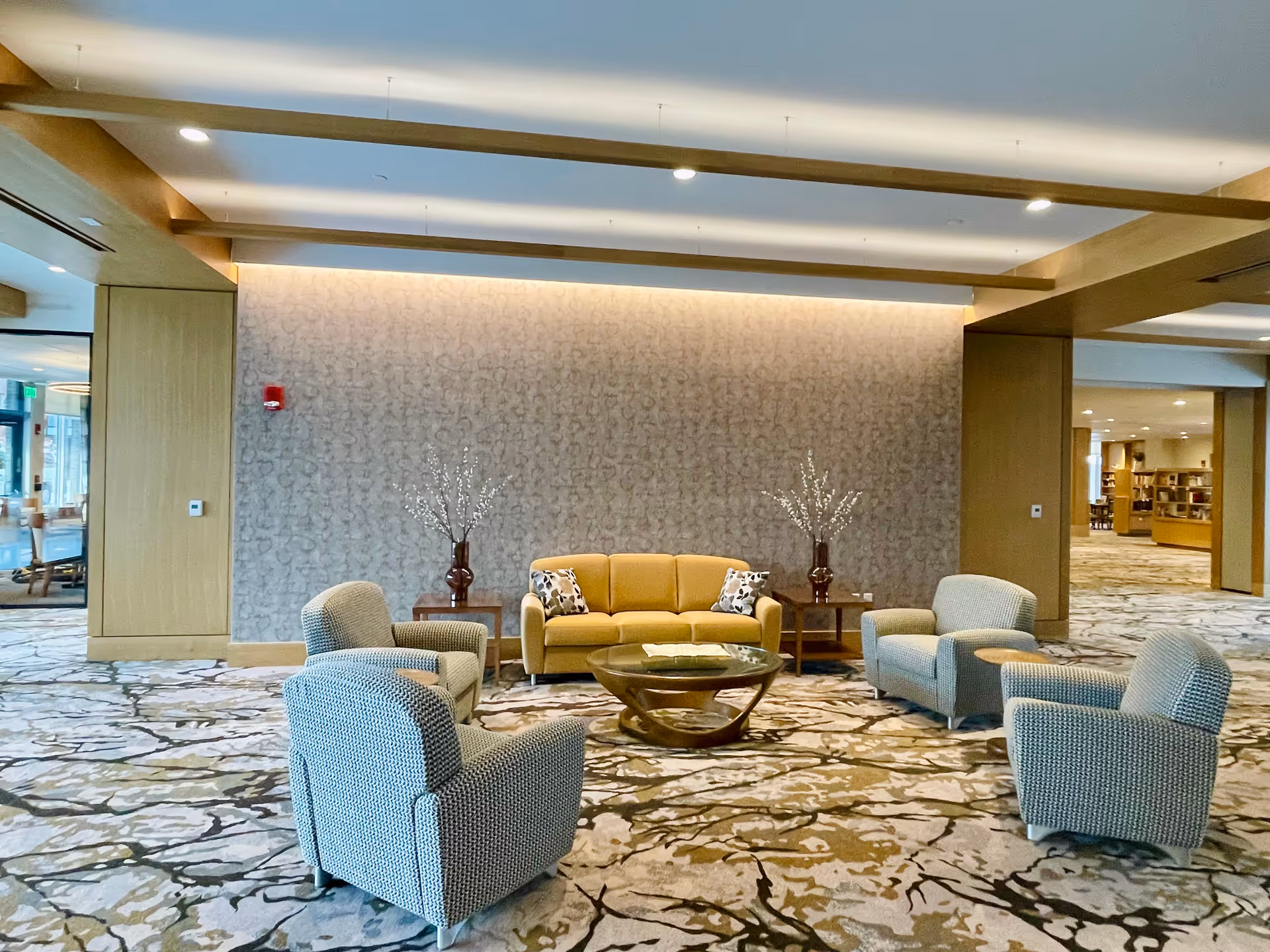 A spacious and well-lit senior living facility lounge area with a patterned carpet, a mustard yellow sofa with two decorative pillows, four gray armchairs arranged around a modern glass coffee table, and two side tables each holding a vase with decorative branches. The walls have a textured design, and wooden beams are visible on the ceiling. There is an open doorway leading to another room with bookshelves in the background.