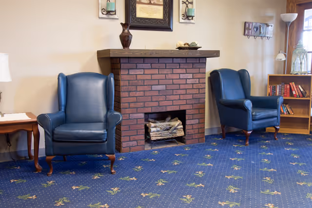 A cozy living area with two blue leather armchairs positioned on either side of a brick fireplace. The fireplace has logs inside and a wooden mantel with decorative items on top. The room features a blue carpet with a floral pattern, a wooden side table with a lamp, a bookshelf with books, and wall decorations including a framed picture and a 'HOME' sign.