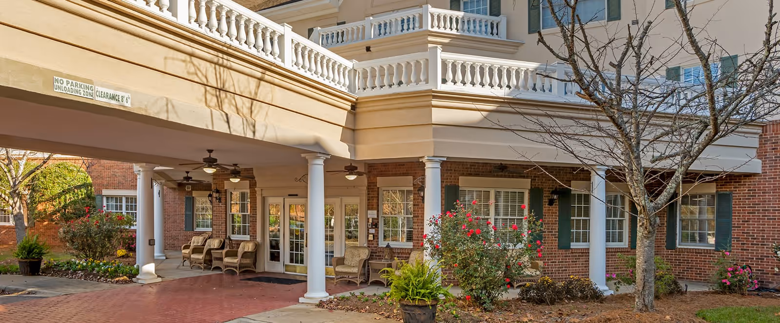 Entrance area of a senior living facility with a covered drop-off zone supported by white columns. There are several wicker chairs arranged under the covered area, ceiling fans, and a brick pathway leading to glass doors. The building exterior is made of red brick with white trim and green shutters. There are landscaped bushes and a tree without leaves in the foreground.