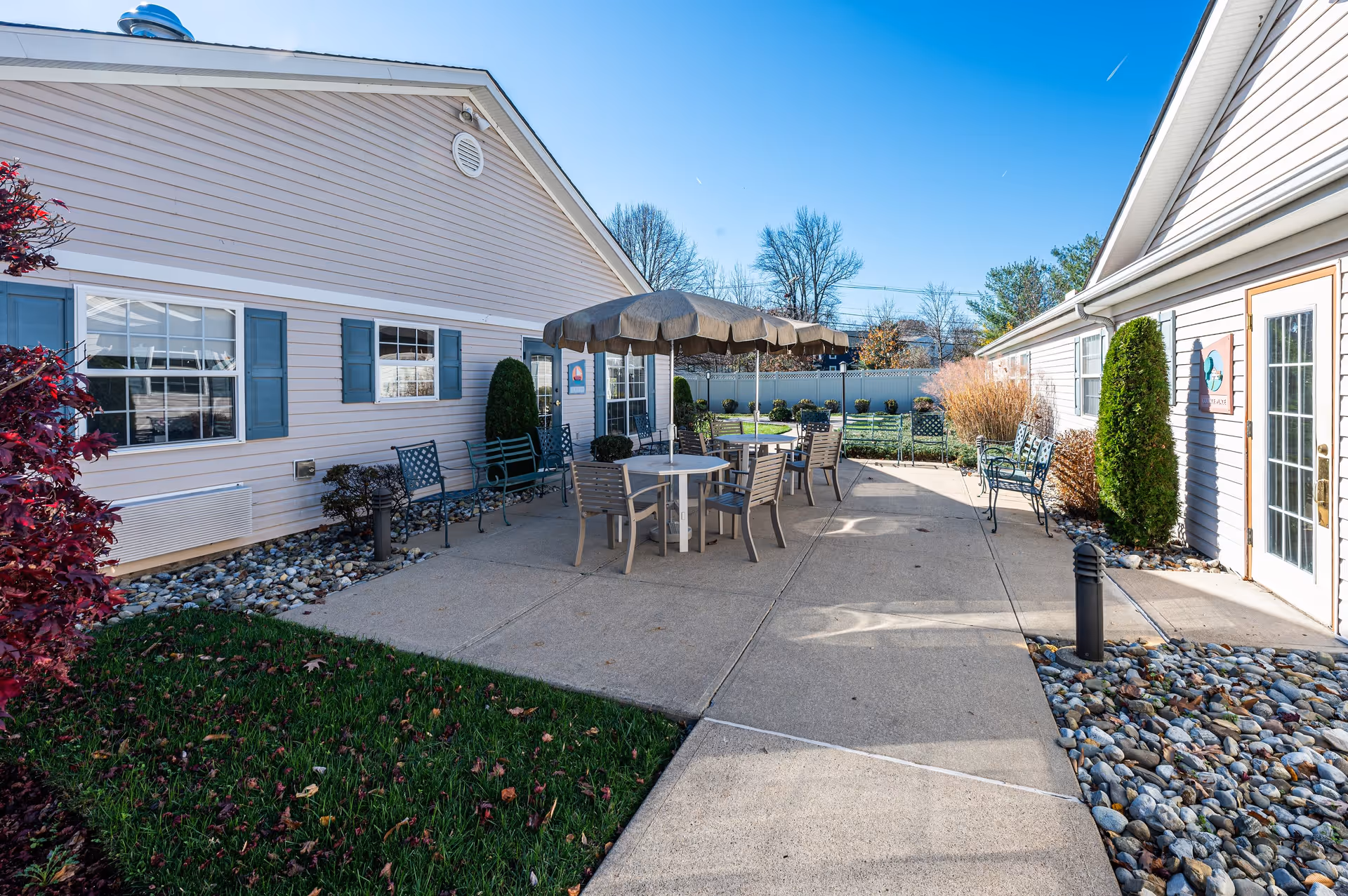 Outdoor patio area between two buildings with several chairs and tables, including one table with a large umbrella. The patio is surrounded by landscaping with bushes, rocks, and grass under a clear blue sky.
