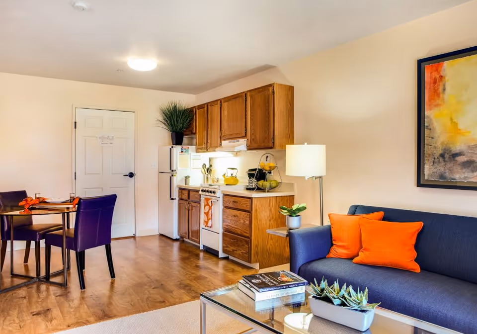 A cozy living space featuring a small kitchen with wooden cabinets, a white refrigerator, and a stove. Adjacent to the kitchen is a dining area with a round glass table and two purple chairs. A blue sofa with two bright orange pillows sits next to a floor lamp and a framed abstract painting on the wall. A glass coffee table in front of the sofa holds a small plant and a few books. The room has wooden flooring and light-colored walls.