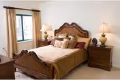 A neatly made bed with a carved wooden headboard and footboard in a bedroom. The bed has a beige bedspread and multiple pillows. There are two matching wooden nightstands on either side of the bed, each with a lamp. A window with beige curtains is visible on the left wall.