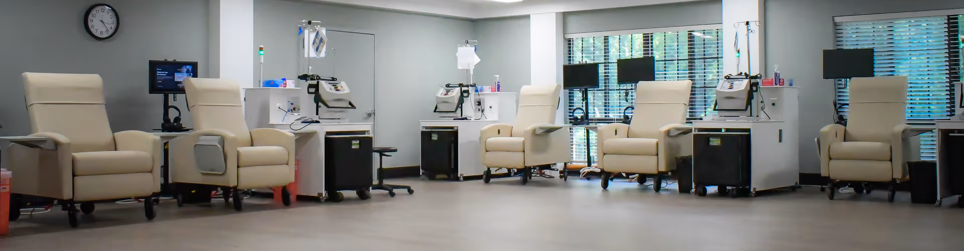 A clinical treatment room with six beige recliner chairs spaced evenly along the walls. Each chair is paired with medical equipment including IV stands and monitors. The room has large windows with blinds, a clock on the wall, and a clean, spacious floor.