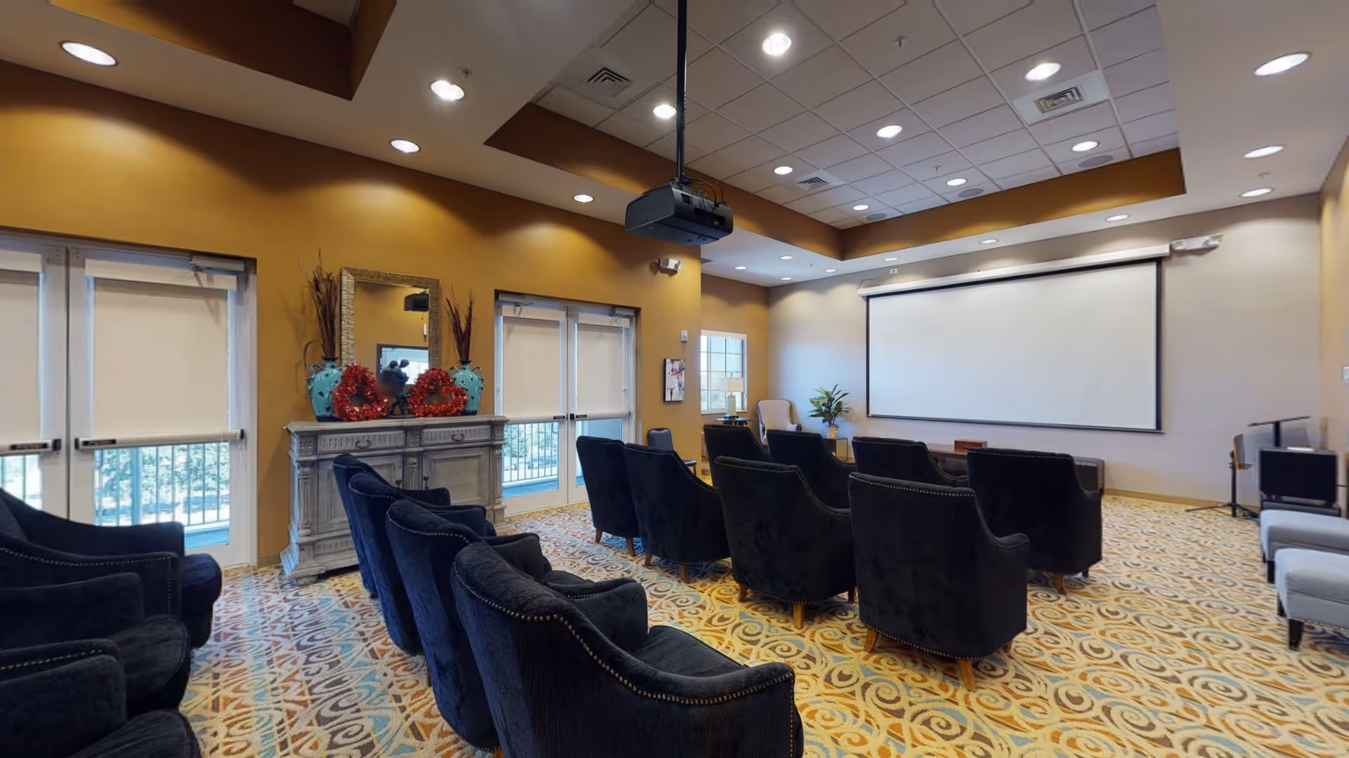 A cozy presentation or movie room with rows of dark upholstered armchairs facing a large white projection screen. The room has a patterned carpet, warm yellow and beige walls, recessed ceiling lights, and a ceiling-mounted projector. There is a decorative cabinet with a mirror and vases on the left side near double doors with window blinds.