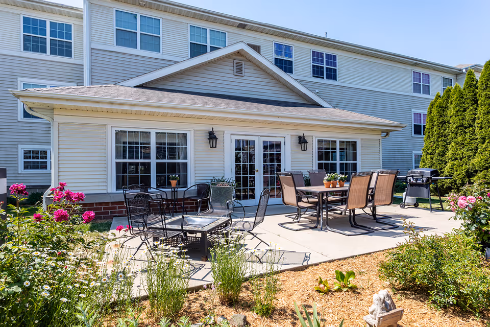 Outdoor patio area at Killarney Kourt with two sets of tables and chairs, surrounded by flowering plants and greenery. The building exterior is light-colored with multiple windows and a double door leading to the patio. A barbecue grill is visible on the right side.
