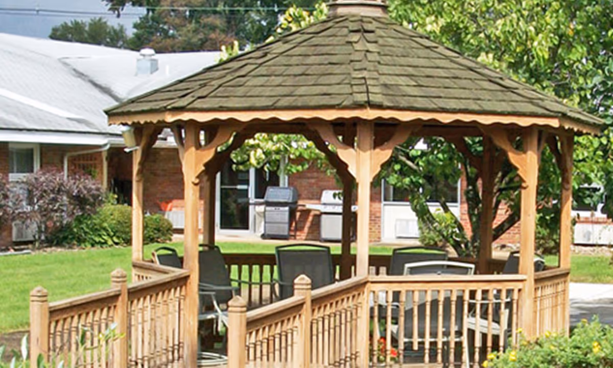 A wooden gazebo with a shingled roof and railing, surrounded by green grass and bushes. Several chairs are placed inside the gazebo. In the background, there is a brick building with windows and a door, along with outdoor grills and trees.