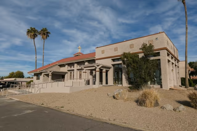 Exterior view of a single-story building with a beige stucco finish and a red-tiled roof, featuring a ramp and steps leading to the entrance. There are two tall palm trees on the left and right sides, with a clear blue sky and some clouds in the background. The surrounding landscape includes gravel and sparse desert plants.