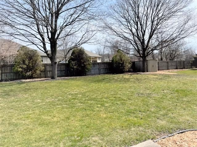 A grassy fenced backyard with leafless trees, shrubs, and a small storage shed under a clear sky.