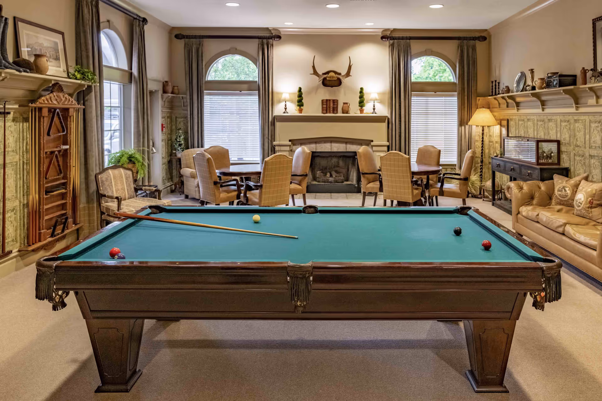 A cozy game room featuring a pool table with balls and a cue stick on it. Behind the pool table, there are several round tables with cushioned chairs arranged around them. The room has large arched windows with curtains, a fireplace with decorative items on the mantel, and a mounted antler decoration above it. To the right, there is a leather sofa with pillows and a floor lamp, and shelves with various decorative objects line the walls.
