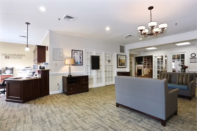 A spacious and well-lit common area in Oak Woods Manor featuring a reception desk with office equipment, two gray upholstered sofas with striped cushions, a wooden side table with a lamp, and glass-paneled double doors leading to another room with bookshelves and a fireplace.