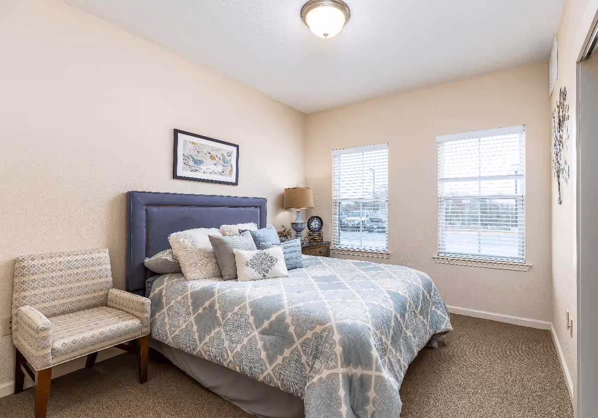 A bedroom with a bed featuring a blue and white patterned comforter and multiple pillows. There is a cushioned chair with wooden legs to the left of the bed, a nightstand with a lamp and clock to the right, and two windows with blinds letting in natural light. A framed picture hangs on the wall above the bed.