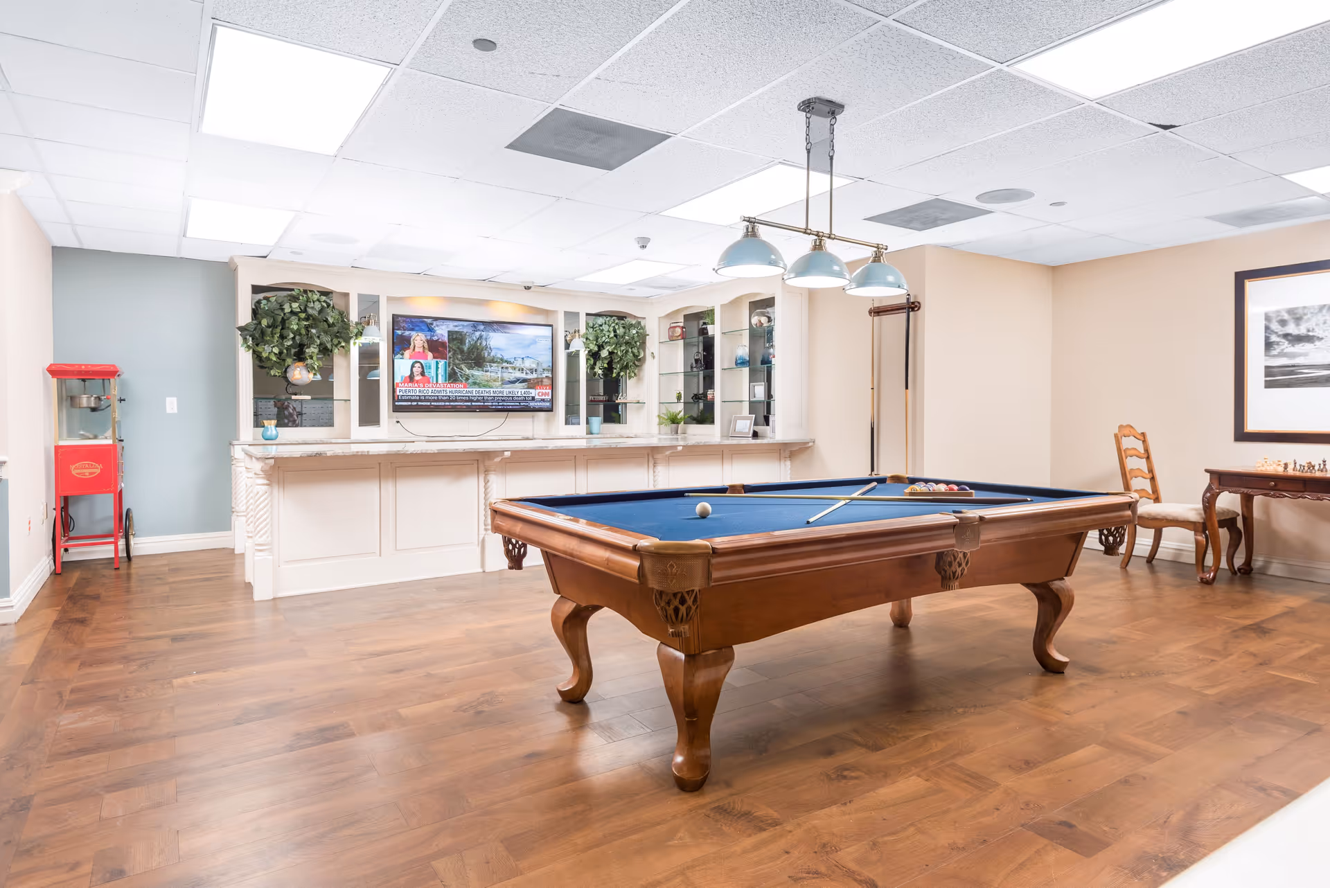 A recreational room with a wooden pool table featuring a blue felt surface, a cue stick, and billiard balls arranged on it. Behind the pool table is a white built-in cabinet with shelves holding plants and decorative items, and a flat-screen TV mounted on the wall displaying a news channel. To the left, there is a red popcorn machine, and to the right, a small wooden table with a chessboard and a wooden chair. The room has wooden flooring, light-colored walls, and a ceiling with recessed lighting and a hanging light fixture above the pool table.