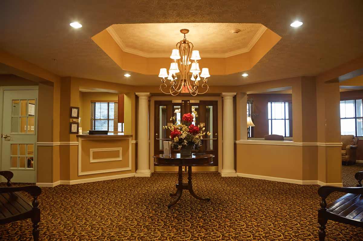 Interior view of a senior living facility lobby area with a round wooden table in the center holding a floral arrangement. The room features beige walls, patterned carpet, a chandelier hanging from a recessed ceiling, and large windows letting in natural light. There are two white columns framing double glass doors at the back, and seating areas are visible on the sides.