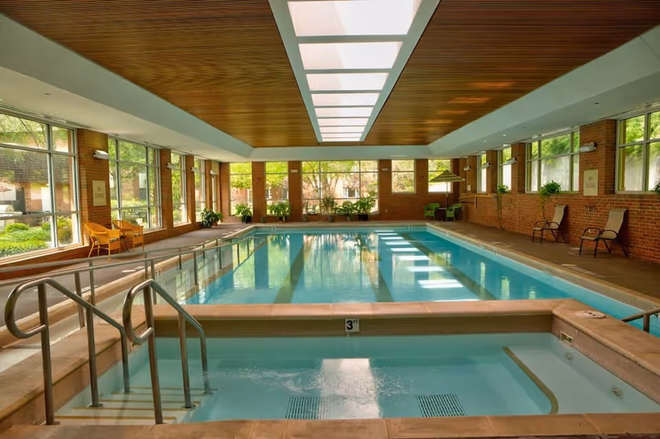 Indoor swimming pool area with a smaller shallow pool in the foreground and a larger pool in the background. The room has large windows on both sides letting in natural light, brick walls, a wooden ceiling with skylights, and several chairs and plants around the pool area.