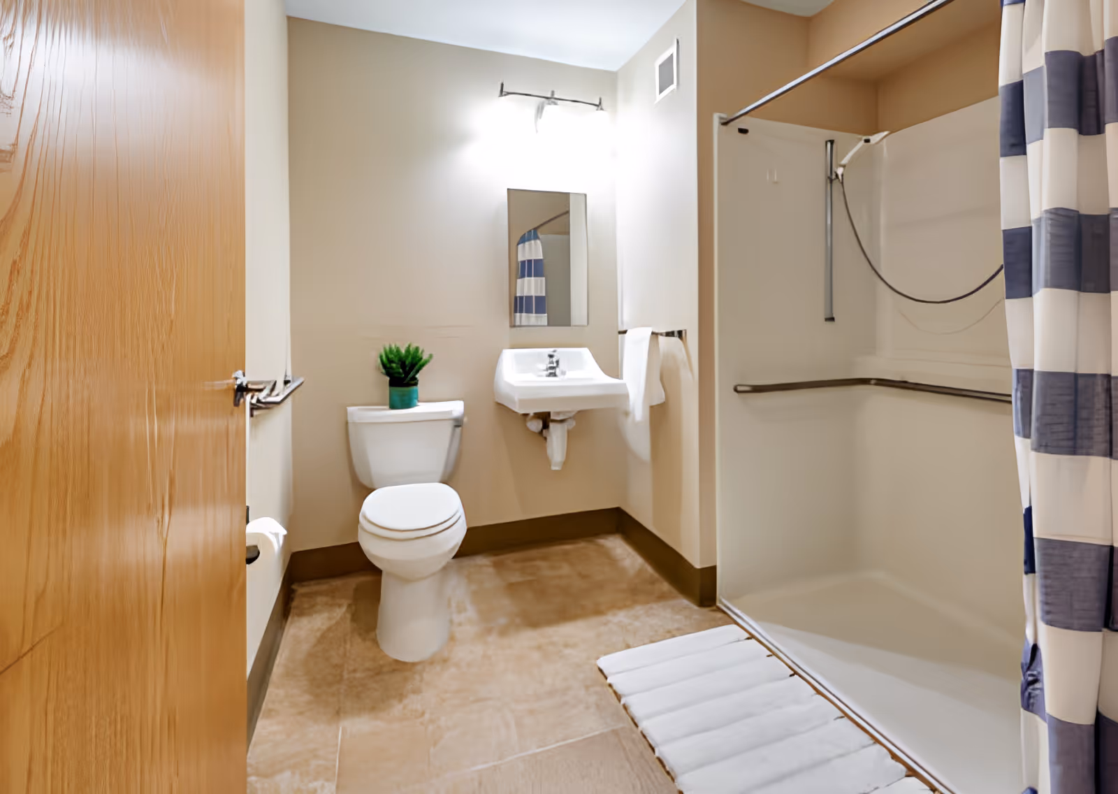 A clean bathroom with a toilet, a small sink with a mirror above it, a shower area with a striped shower curtain, and a small green plant on the toilet tank. The bathroom has beige walls and a light brown tiled floor.