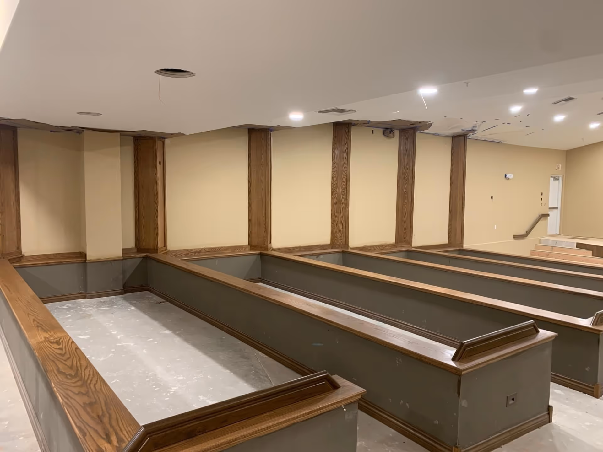 Interior view of a room under renovation with multiple wooden framed planter boxes or raised garden beds along the walls and floor. The ceiling has visible damage with holes and missing panels, and the floor is unfinished. The walls are painted beige with wooden trim accents.