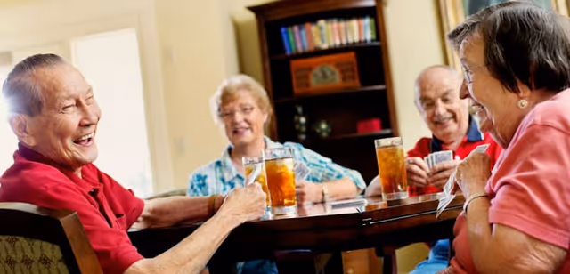 Four elderly people sitting around a round wooden table playing cards and enjoying drinks in a cozy indoor setting with a bookshelf in the background.