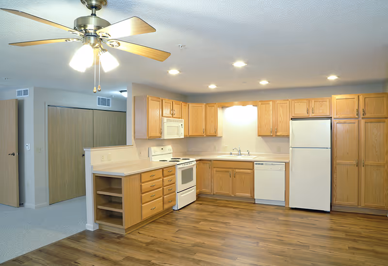 A clean and spacious kitchen with wooden cabinets, white appliances including a refrigerator, stove, microwave, and dishwasher. The kitchen has a wooden floor and a ceiling fan with lights. Adjacent to the kitchen is a carpeted area with a closed wooden door and closet doors.