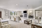 A spacious and well-lit living room with multiple upholstered chairs and sofas arranged around a central coffee table. The room features a fireplace with a mounted flat-screen TV above it, large windows allowing natural light, and neutral-colored walls and carpet. Decorative framed pictures and lamps add to the cozy ambiance.