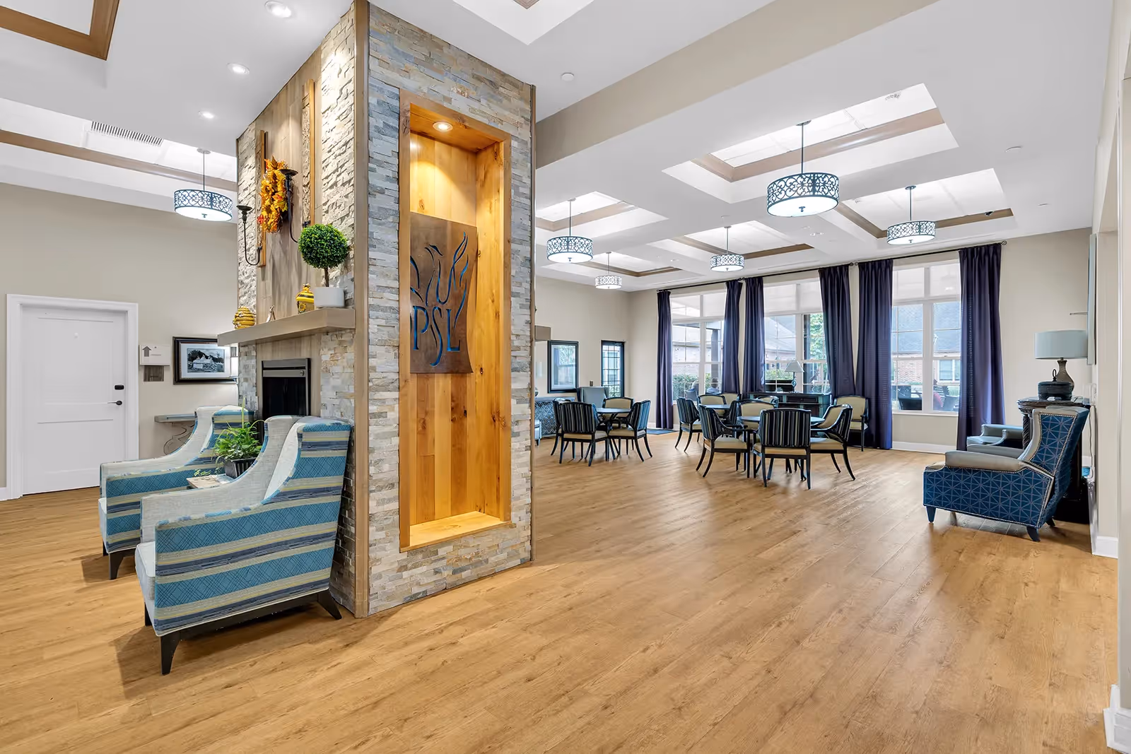 Bright, open memory care community common room with seating areas, dining tables, a decorative fireplace feature, and large windows.
