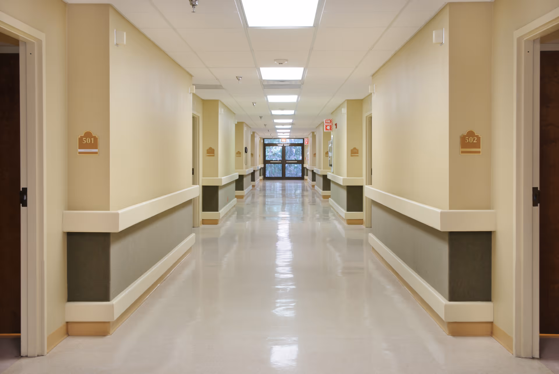 Empty, well-lit hallway in a senior living facility with room numbers (501, 502), handrails along the walls, and an exit at the far end.