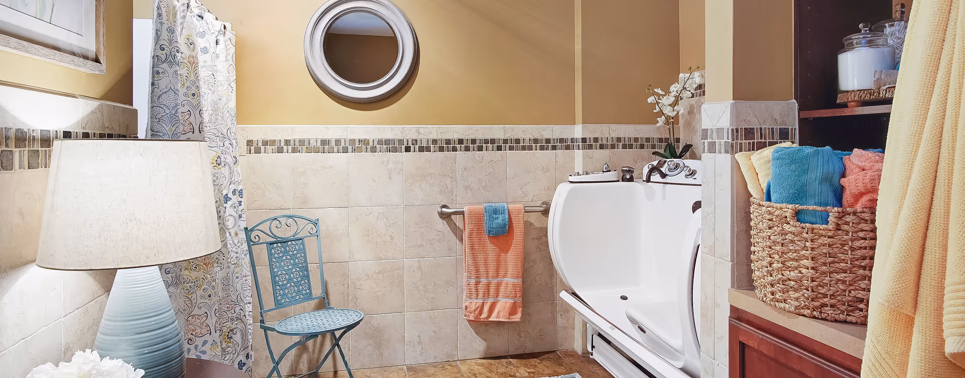 A bathroom with beige tiled walls and floor, featuring a white walk-in bathtub with controls on the side. There is a blue metal chair, a lamp with a blue base and beige shade, a round mirror on the wall, and a basket filled with colorful towels on a wooden shelf. A towel rack holds an orange towel with a smaller blue towel on top. A floral-patterned shower curtain is partially visible.