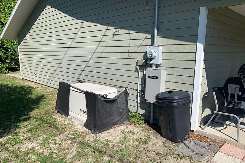 Outdoor side view of a building with light green siding, showing an electrical meter and panel mounted on the wall, a large generator partially covered with a black mesh screen, a black trash bin, and two chairs on a small concrete patio area.