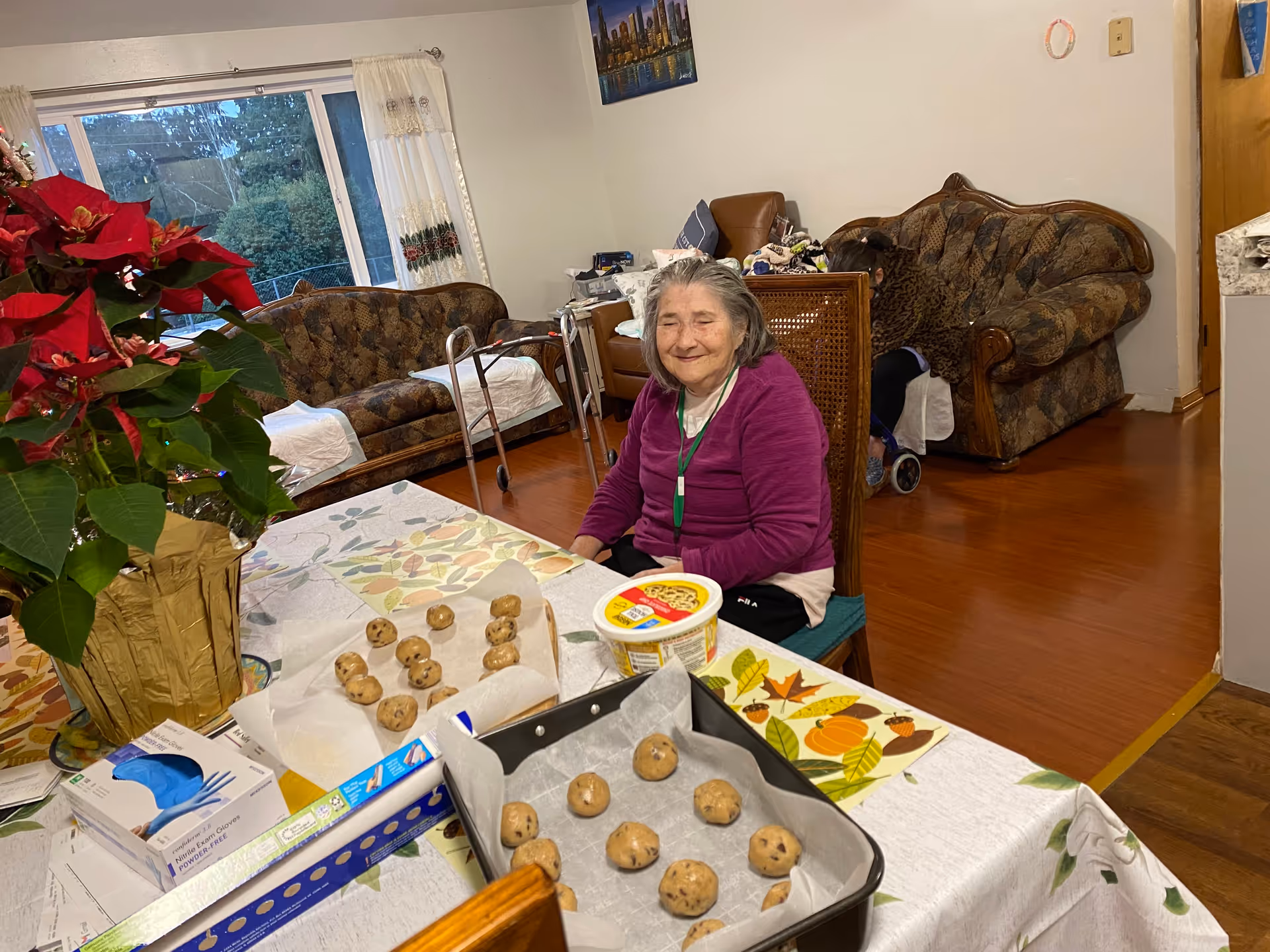 An elderly woman wearing a purple sweater is sitting at a dining table with cookie dough balls on parchment paper and in a baking tray. The table is decorated with a poinsettia plant and a fall-themed placemat. In the background, there are two patterned sofas, a walker, and another person sitting on one of the sofas. The room has wooden flooring and a large window with white curtains.