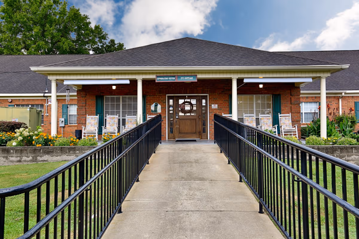 Front entrance of Country Cottage Montgomery facility with a concrete ramp and black metal railings leading to a wooden door. The building is made of red brick with white columns supporting a covered porch. There are several rocking chairs with floral cushions on the porch and flower beds with colorful flowers on either side of the ramp. The sky is partly cloudy.