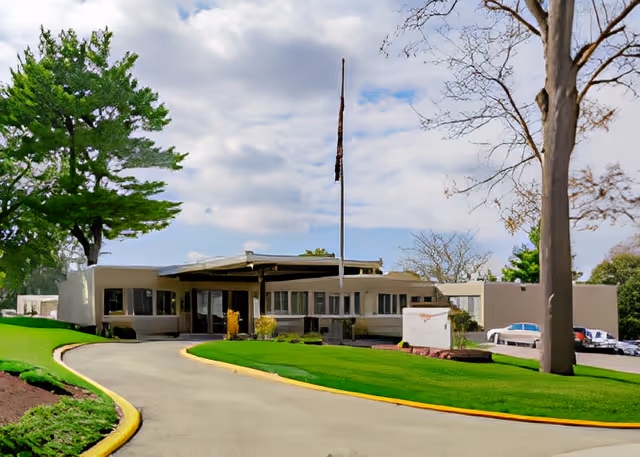 Exterior view of a single-story senior living facility building with a curved driveway leading to the entrance. The building is surrounded by well-maintained green lawns, trees, and a flagpole in front. Several parked cars are visible on the right side of the building under a partly cloudy sky.